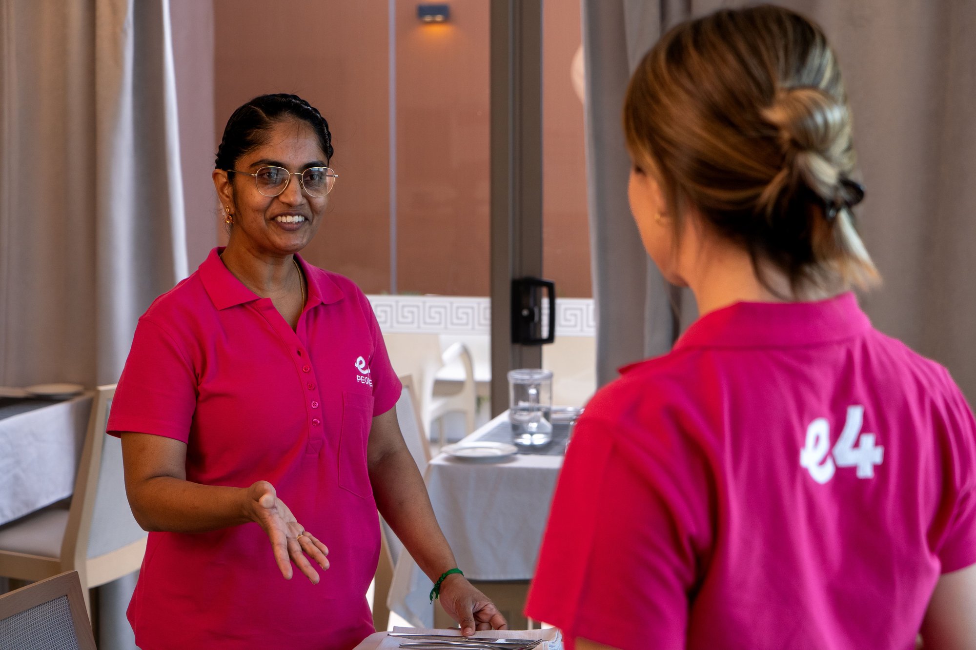 Two E4 nurses, back shot of one with E4 logo on back on pink uniform, talking to eachother in dining room 4